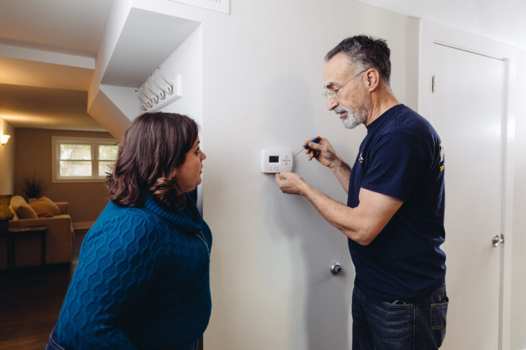 A Fixer working on a thermostat while a homeowner watches